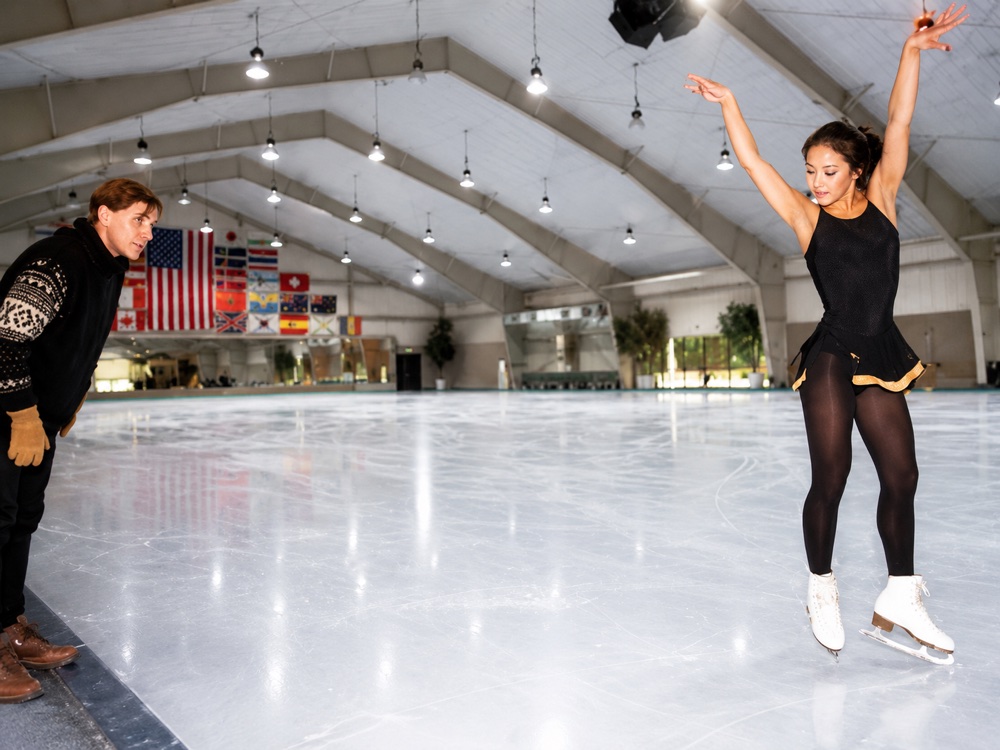 Figure skater Michelle Kwan mid-jump on the ice at the mountain arena, her coach watching from the boards